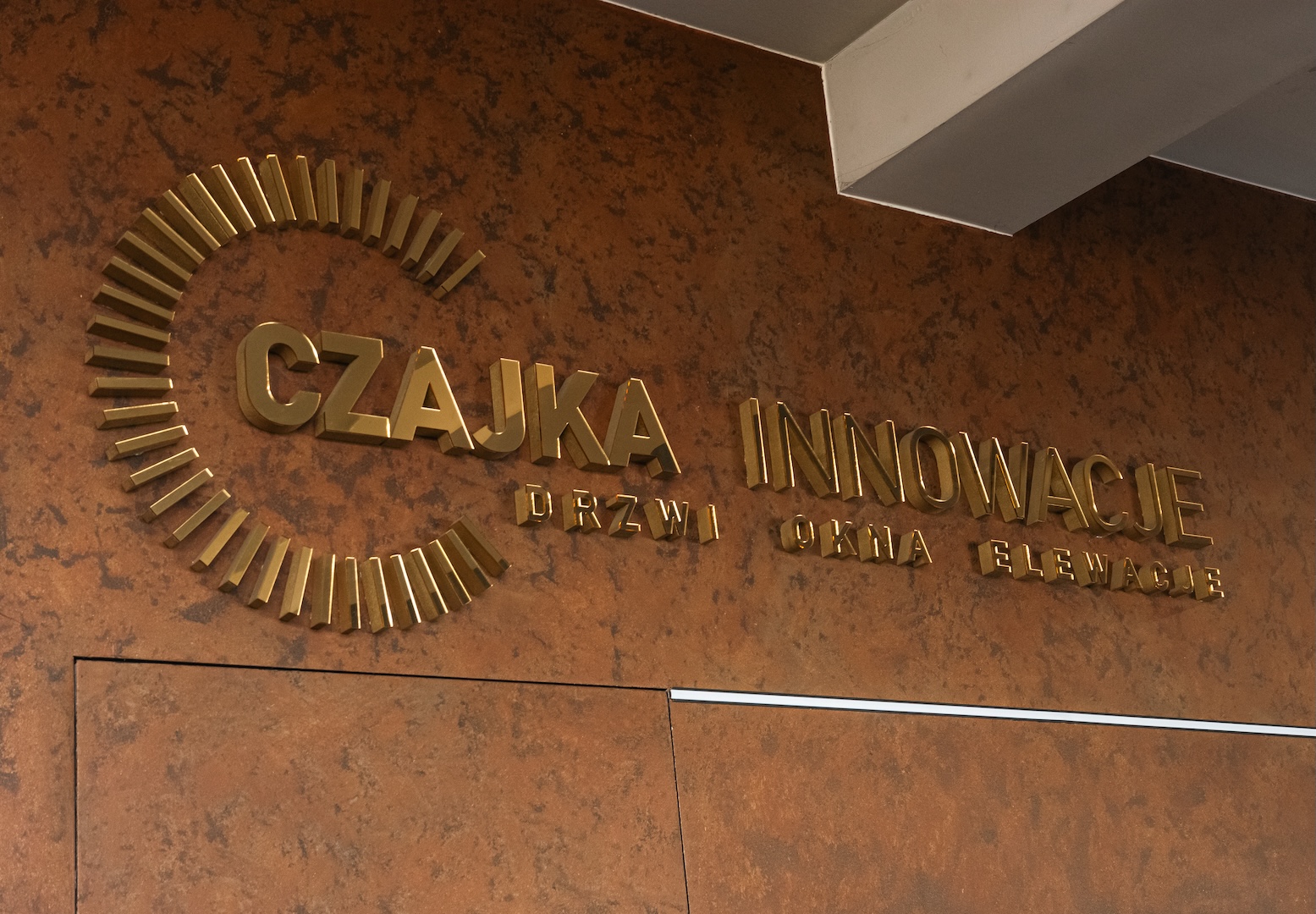 Mosiężny logotyp Czajka Innowacje na fakturowanej miedzianej ścianie salonu